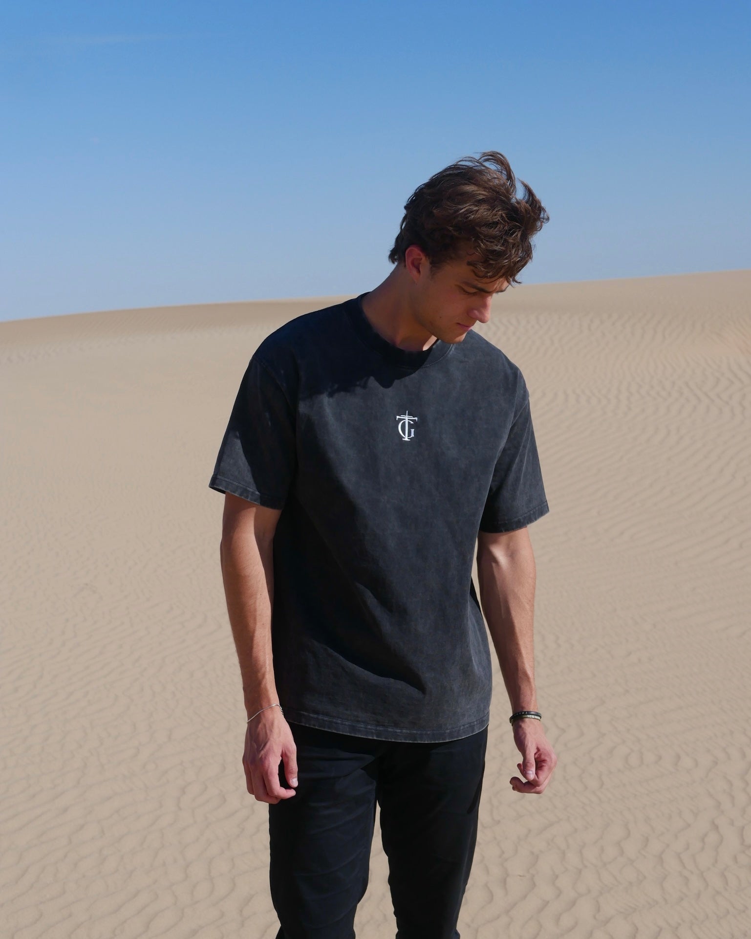 Model wearing black sanded Thank God T-shirt with TG cross logo, standing in sunlit desert landscape – Christian streetwear tee made from heavyweight washed cotton, blending faith and fashion