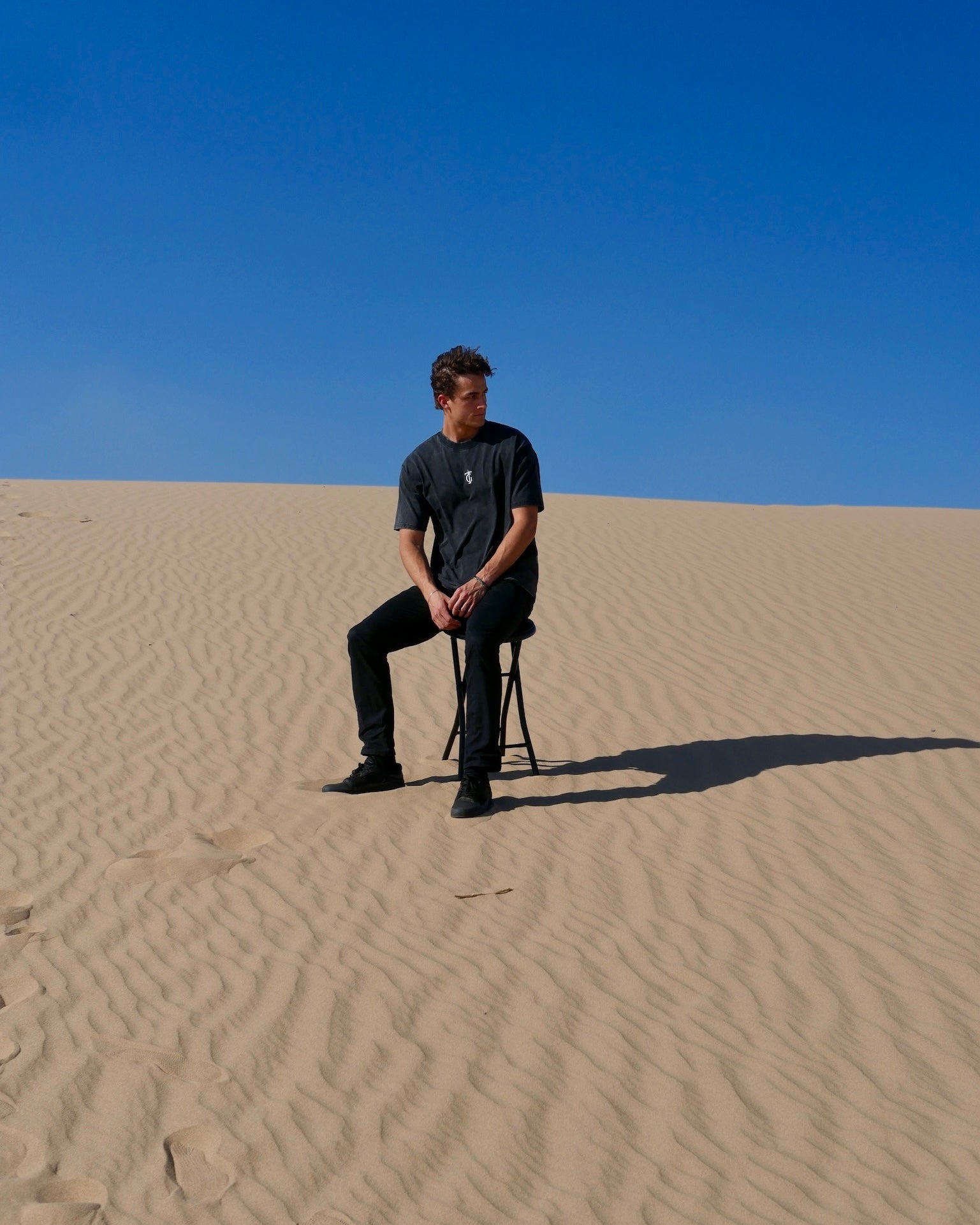 Model wearing black sanded Thank God T-shirt with TG cross logo, sitting in sunlit desert landscape – Christian streetwear tee made from heavyweight washed cotton, blending faith and fashion.